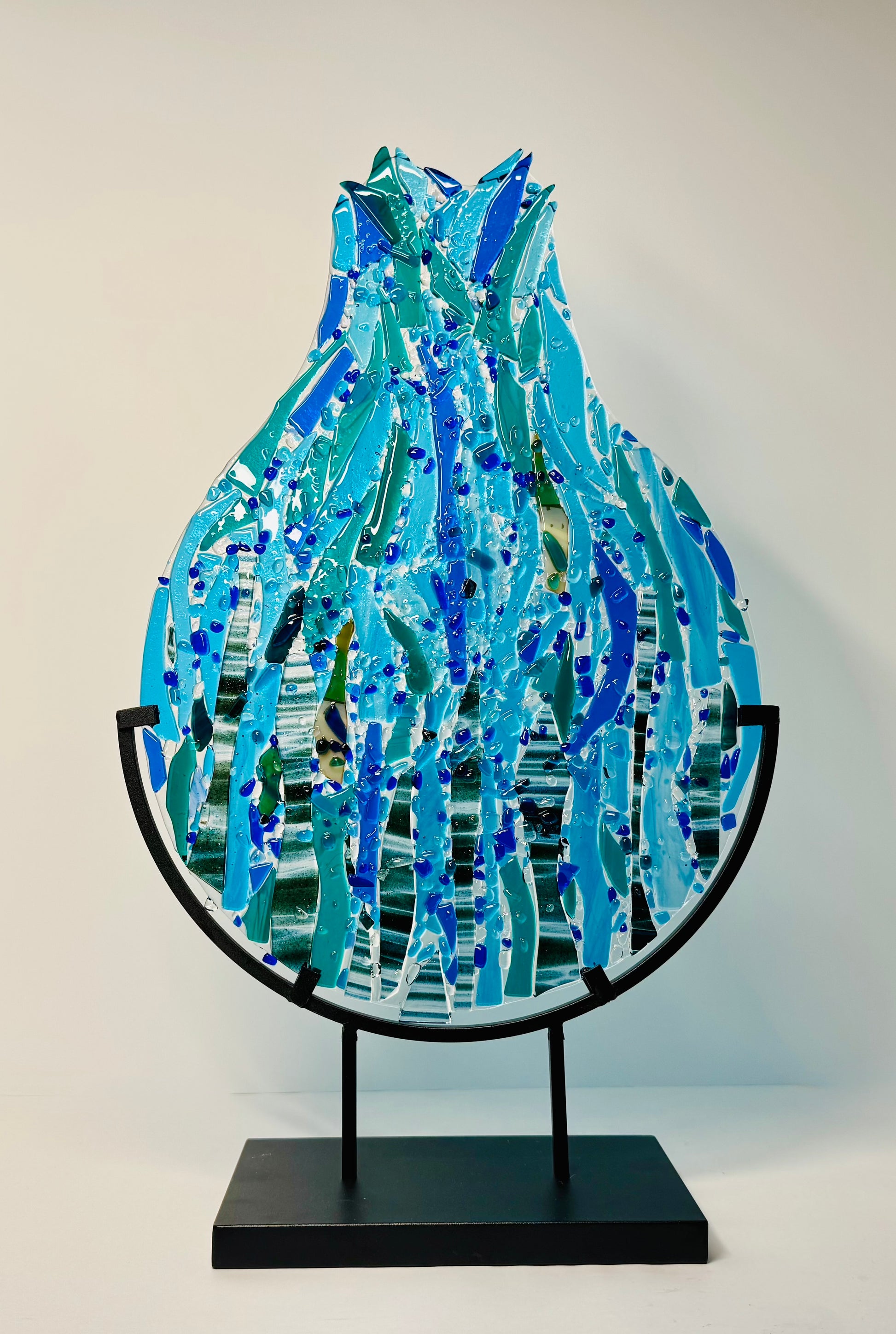 Decorative abstract glass sculpture to represent a teardrop or raindrop water design of various blues in a black metal stand.